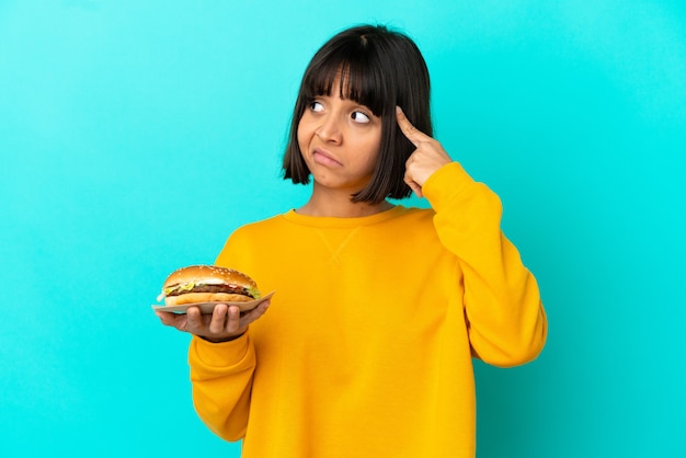 Foto mujer joven morenita sosteniendo una hamburguesa sobre antecedentes aislados teniendo dudas y pensando