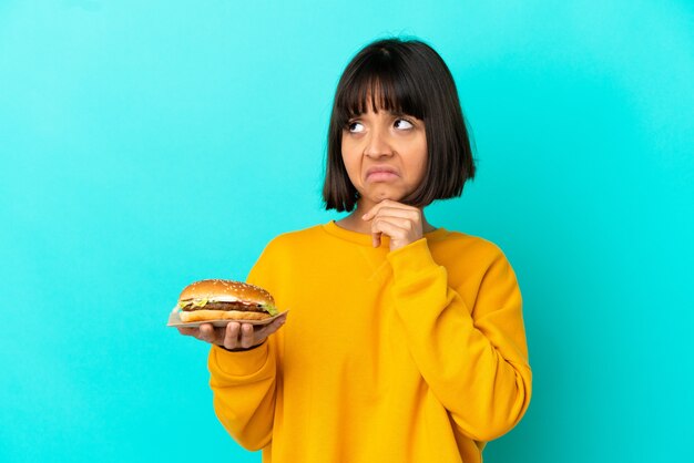 Mujer joven morenita sosteniendo una hamburguesa sobre antecedentes aislados teniendo dudas