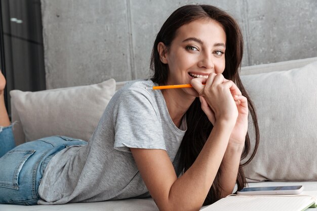 Mujer joven morenita vistiendo ropa casual relajándose en un sofá, estudiando, sosteniendo un lápiz