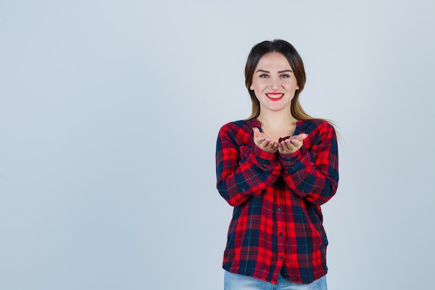 Mujer joven mostrando recibir o dar gesto en camisa a cuadros y mirando alegre. vista frontal.