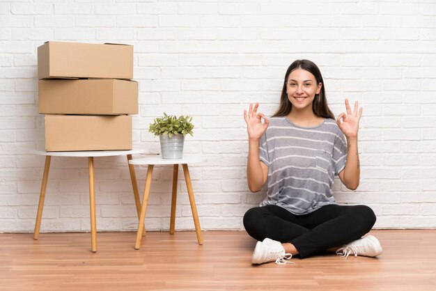 Mujer joven moviéndose en un nuevo hogar entre cajas que muestran un signo bien con los dedos