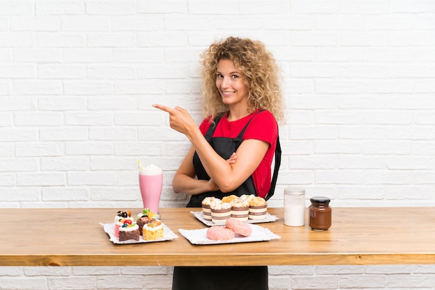 Mujer joven con muchos mini pasteles diferentes en una mesa, apuntando con el dedo hacia un lado