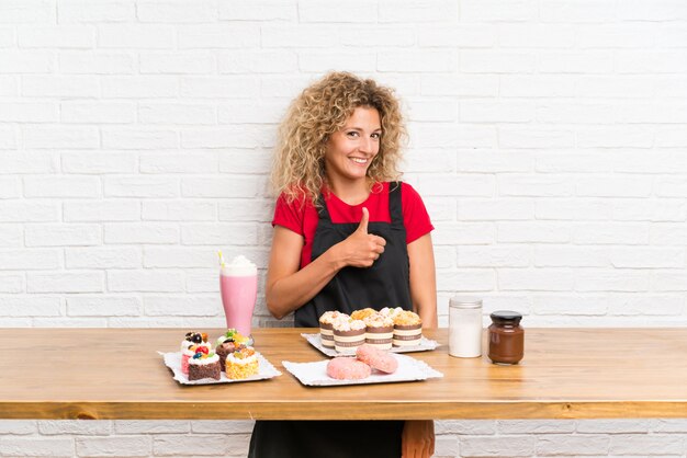 Mujer joven con muchos mini pasteles diferentes en una mesa dando un gesto de aprobación