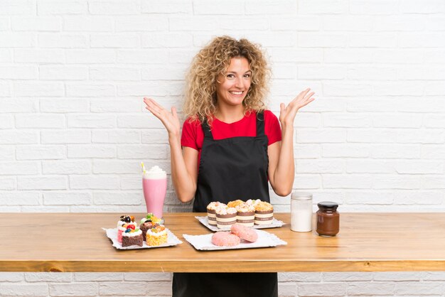 Mujer joven con muchos mini pasteles diferentes en una mesa con expresión facial sorpresa