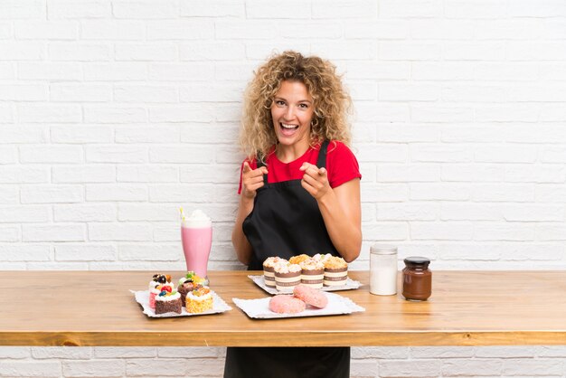 Mujer joven con muchos mini pasteles diferentes en una mesa te señala con el dedo