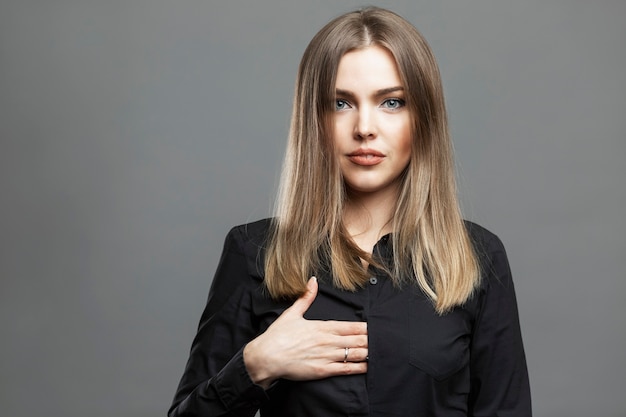 Mujer joven muestra una señal de mano oculta. Hermosa rubia con una camisa negra. El simbolismo masónico y la teoría de la conspiración mundial. Fondo gris.