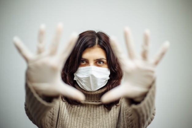 Mujer joven muestra signos con las manos y tiene que quedarse en casa durante la cuarentena debido a la pandemia de coronavirus. Hermosa chica sentada en su piso con guantes y máscara médica. Concepto de epidemia de Covid-19.