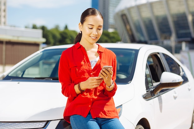 Mujer joven. Mujer positiva inteligente apoyado en el coche mientras mira la pantalla de su teléfono inteligente