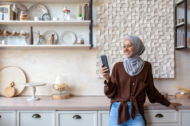 Mujer joven musulmana sonriente con hiyab parada en la cocina de casa y usando el teléfono móvil leyendo noticias