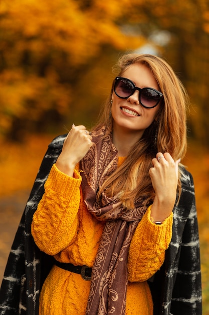 Foto mujer joven muy feliz con linda sonrisa en ropa de moda de otoño: suéter, abrigo y gafas de sol disfrutan y camina al aire libre con coloridas hojas amarillas increíbles