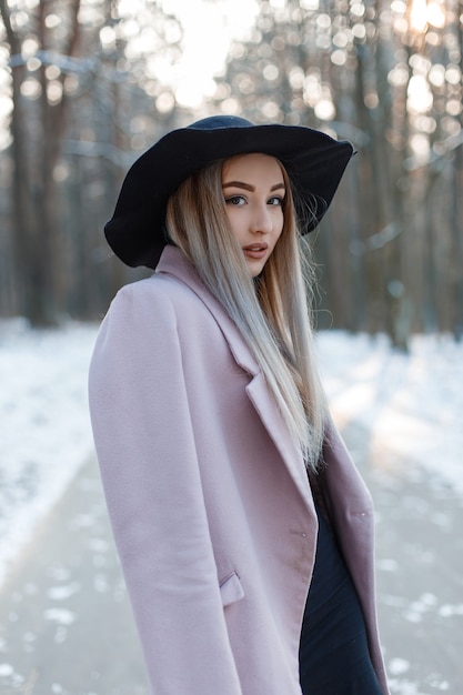 Mujer joven muy hermosa en ropa de glamour de invierno con estilo va con un sombrero elegante en un bosque nevado en un día soleado de invierno. Chica elegante atractiva de moda.