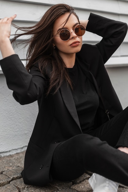 Mujer joven muy de moda en gafas de sol de moda en ropa juvenil negra fresca en zapatillas blancas se relaja en baldosas de piedra cerca de edificio vintage en la ciudad. Chica de moda en traje elegante posa en la calle.