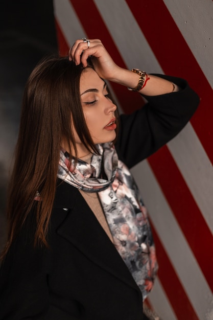 Mujer joven muy sexy en ropa elegante juvenil posando en la ciudad. Modelo de moda chica sensual en abrigo negro con bufanda de primavera de seda de moda se relaja cerca de una columna con línea roja-blanca al aire libre.