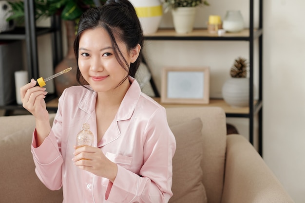 Mujer joven muy sonriente aplicando suero con vitamina c en su rostro antes de irse a la cama