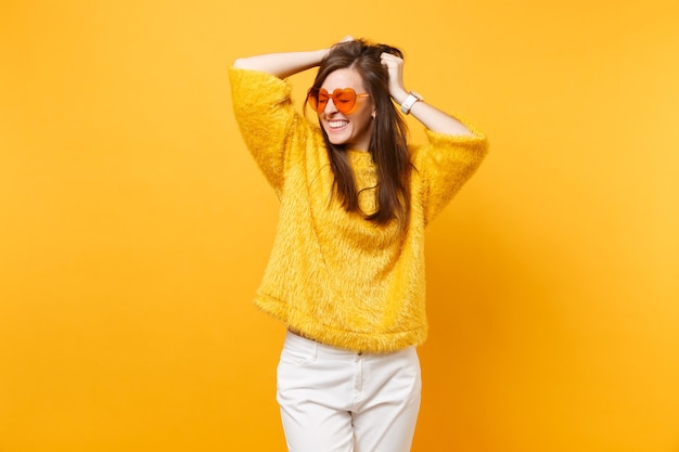 Mujer joven muy sonriente con los ojos cerrados en suéter de piel y gafas de corazón naranja aferrándose a la cabeza aislada sobre fondo amarillo brillante. Personas sinceras emociones, concepto de estilo de vida. Área de publicidad.