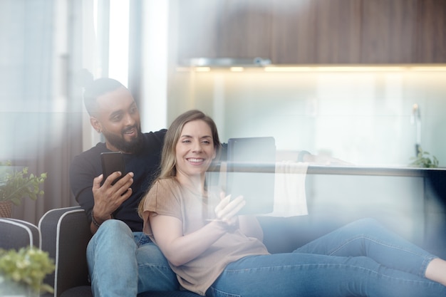 Mujer joven muy sonriente que muestra algo interesante que encontró en Internet al marido cuando están descansando en el sofá en casa