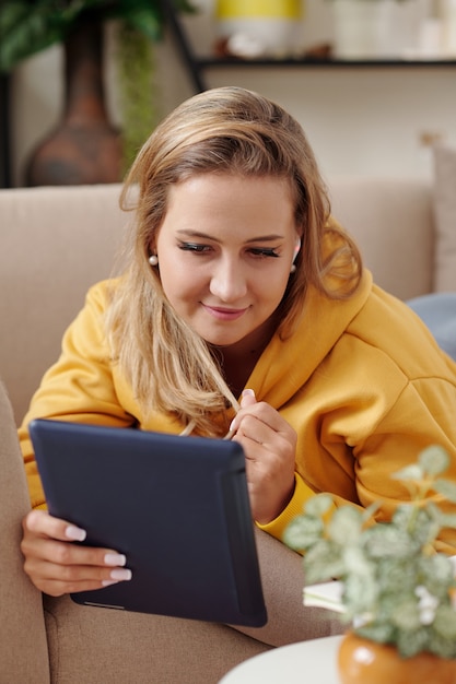 Mujer joven muy sonriente relajándose en el sofá y leyendo un libro electrónico o viendo una película en la computadora de la tableta