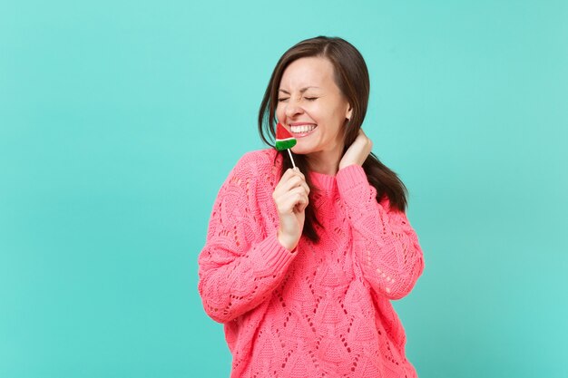 Mujer joven muy sonriente en suéter rosa de punto con los ojos cerrados sosteniendo en la mano piruleta de sandía aislado sobre fondo de pared azul, retrato de estudio. Concepto de estilo de vida de personas. Simulacros de espacio de copia.