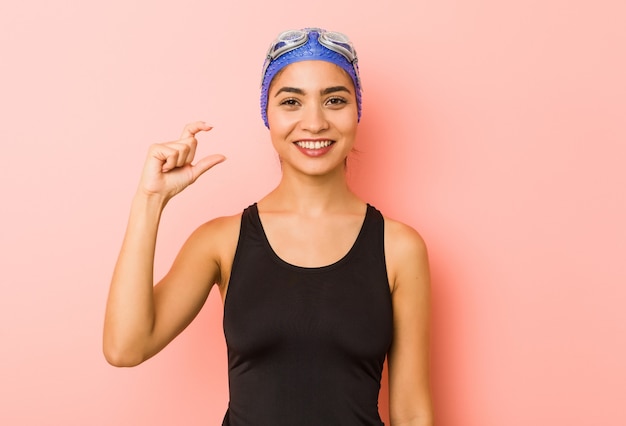 Mujer joven nadadora árabe aislada sosteniendo algo pequeño con los dedos índices, sonriente y confiado.