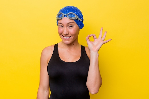 Foto mujer joven nadadora australiana aislada sobre fondo amarillo guiña un ojo y tiene un buen gesto con la mano.