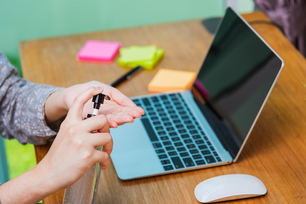 Mujer joven de negocios asiáticos que trabaja desde su oficina en casa, pone en cuarentena el coronavirus de la enfermedad o COVID-19 usando una máscara protectora y limpiando las manos con gel desinfectante en la computadora portátil delantera