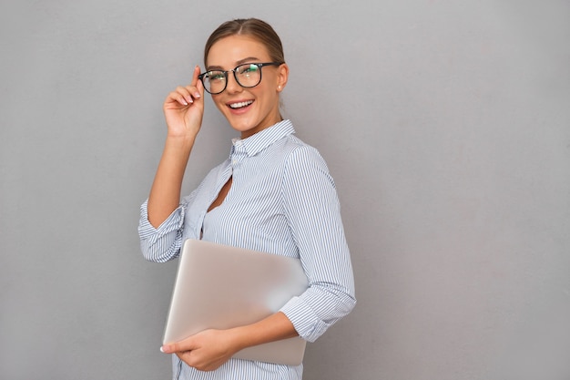 Mujer joven de negocios feliz posando aislada sobre fondo de pared gris con ordenador portátil.