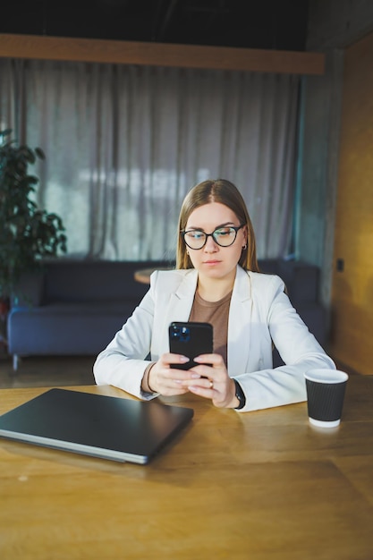 Mujer joven de negocios con gafas y ropa informal sentada en una mesa de madera con una computadora portátil y una taza de café haciendo la tarea en una computadora portátil