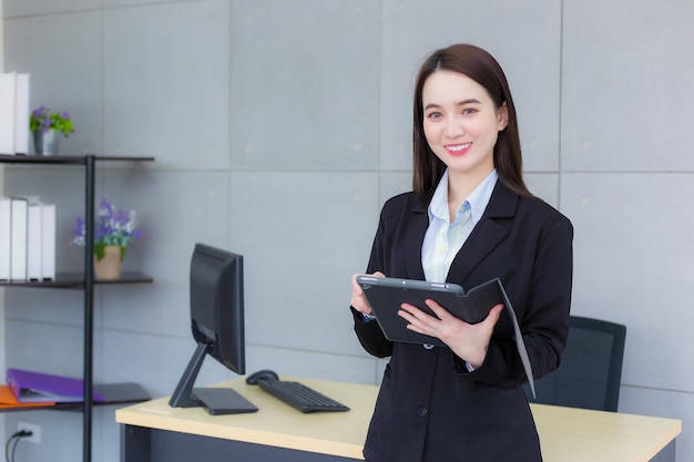 Mujer joven de negocios profesional asiática en traje negro sonríe felizmente de pie y mira a la cámara