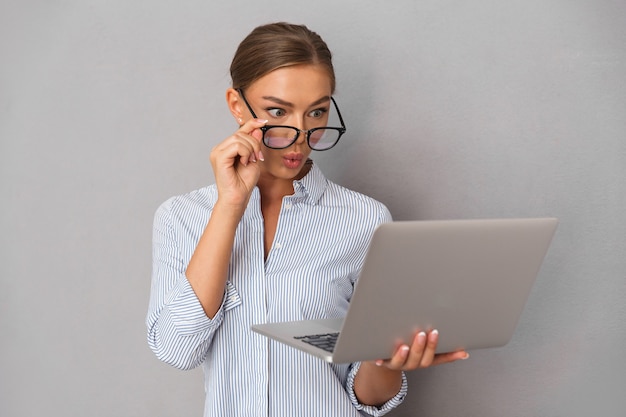 Mujer joven de negocios sorprendida posando aislada sobre fondo gris de la pared usando la computadora portátil.