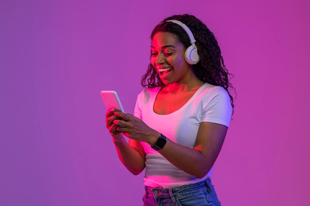 Mujer joven negra emocionada usando auriculares inalámbricos usando un teléfono inteligente en luz de neón