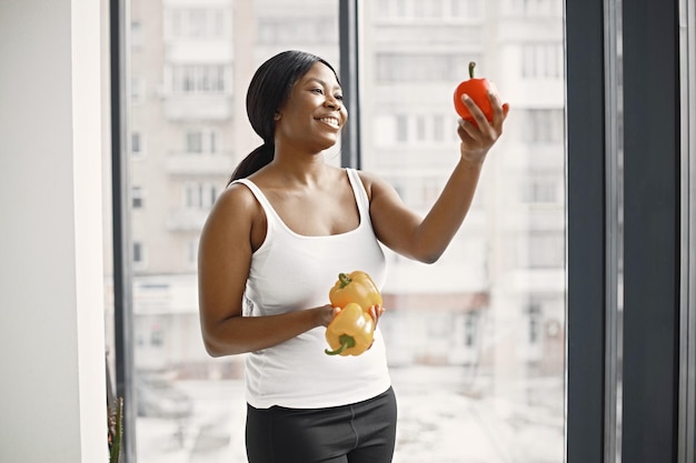 Mujer joven negra en estudio con ventana grande sosteniendo un pimiento amarillo y rojo