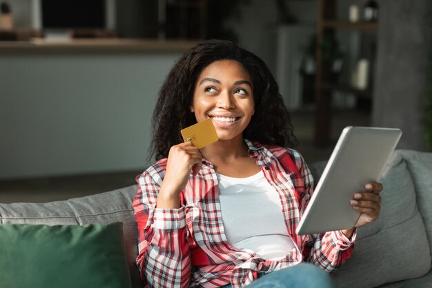 Mujer joven negra soñadora satisfecha con tarjeta de crédito y tableta piensa en comprar disfruta de compras y ventas en línea