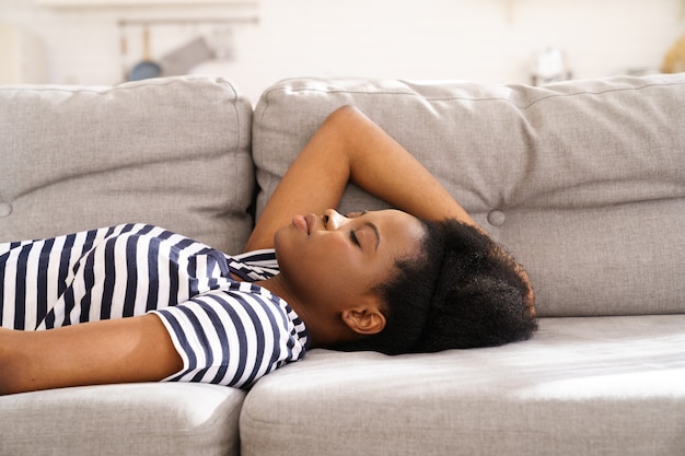 Mujer joven negra usar camiseta despojada descansando durmiendo en el sofá en casa ojos cerrados tomando un descanso