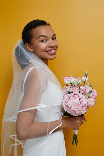 Mujer joven negra con vestido de novia y sosteniendo flores contra amarillo