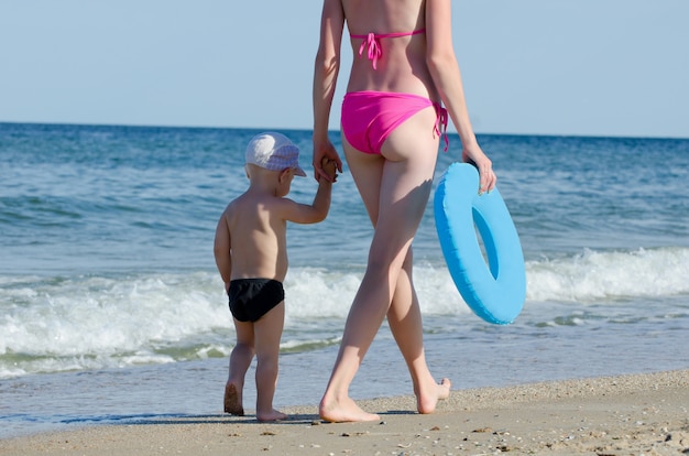 Mujer joven con un niño pequeño ir a lo largo de la línea del mar, día soleado de verano