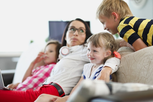 Una mujer joven con niños está sentada en el sofá de la sala de estar y viendo la televisión mamá