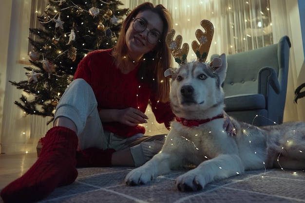 Mujer joven en la noche de Navidad junto con un perro mascota divirtiéndose poniendo cuernos de venado de Navidad y guirnaldas sentados en el suelo hasta el árbol de Navidad mira la cámara