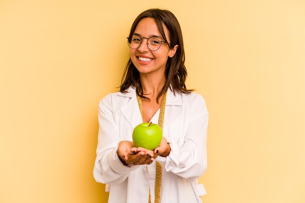 Mujer joven nutricionista sosteniendo una máquina de pesaje aislada sobre fondo amarillo