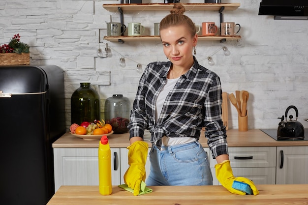 Mujer joven o ama de casa en guantes de goma limpiando la mesa con un paño de microfibra en la cocina de casa.