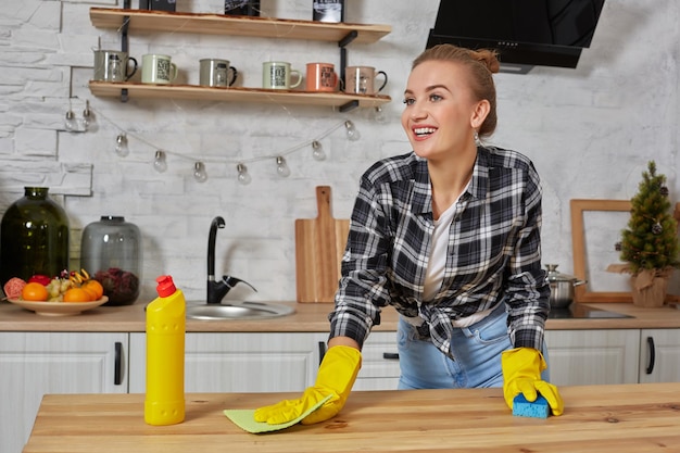 Mujer joven o ama de casa en guantes de goma limpiando la mesa con un paño de microfibra en la cocina de casa.