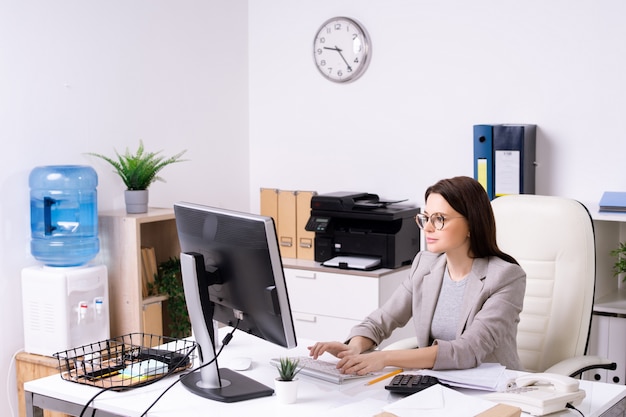 Mujer joven ocupada en gafas escribiendo en el teclado de la computadora mientras trabaja en la oficina moderna