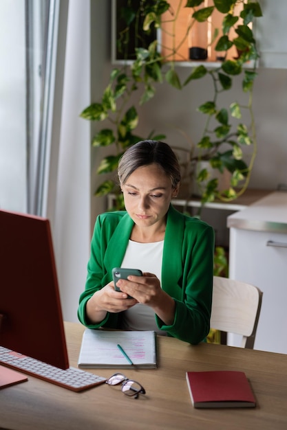 Mujer joven en la oficina en casa usando un teléfono inteligente con redes sociales Lugar de trabajo acogedor con ventana grande