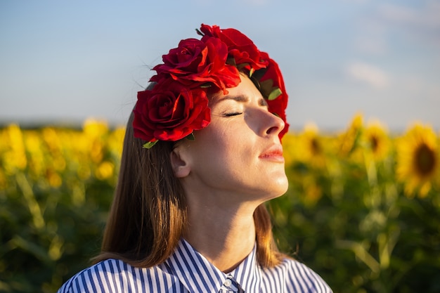 Mujer joven con los ojos cerrados con una corona de flores rojas en un campo de girasoles al atardecer.