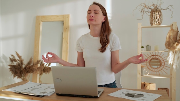 Mujer joven con los ojos cerrados meditación extendió las manos en pose de yoga sentarse después del trabajo en el escritorio con el portátil