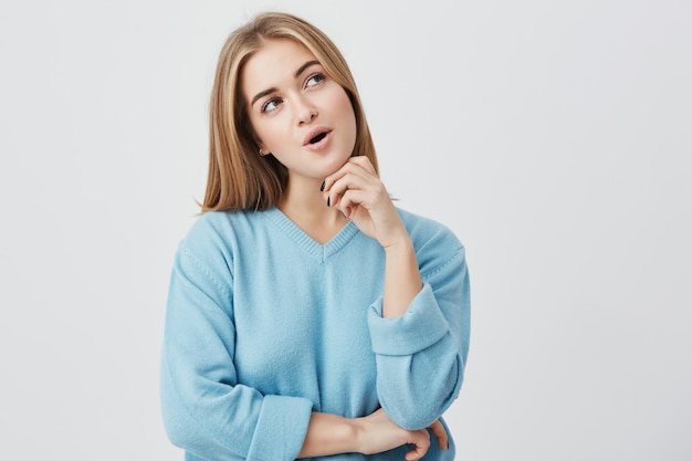 Mujer joven de ojos oscuros con cabello rubio y suéter azul con expresión pensativa pensando en sus planes mirando a un lado