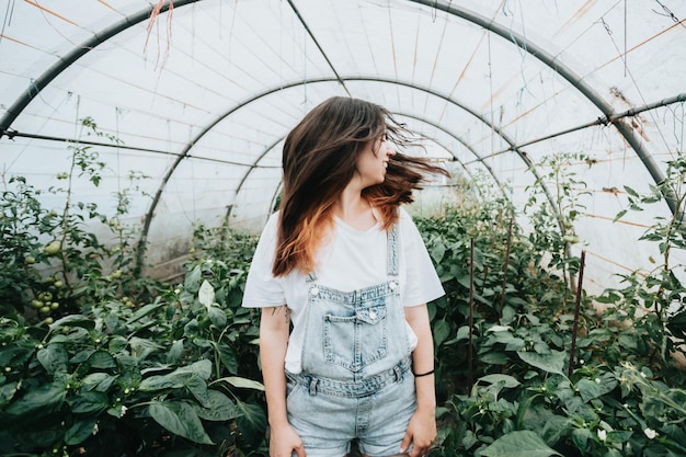 Mujer joven ondeando el cabello de pie en un invernadero cerca de plantas con verduras Sostenibilidad y concepto de crecimiento responsable Cosecha de alimentos ecológicos y biosaludables