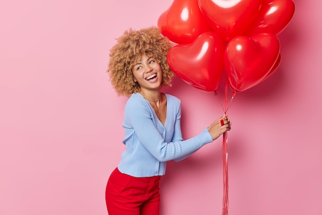 Mujer joven optimista con una amplia sonrisa vestida con un puente casual sostiene un montón de globos inflados en forma de corazón celebra el Día de San Valentín aislado sobre fondo rosa Ocasión festiva