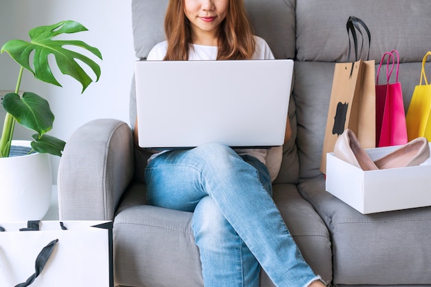 Mujer joven con ordenador portátil sentado en el sofá con sus artículos de moda de compras paquete en línea. Compras en línea, banca por Internet, trabajo en / desde casa, nuevo concepto de estilo de vida normal