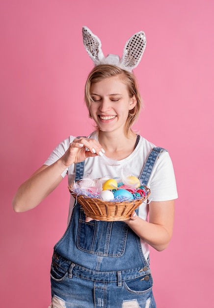 Mujer joven en orejas de conejo con canasta con huevos de Pascua de colores aislados en pared rosa