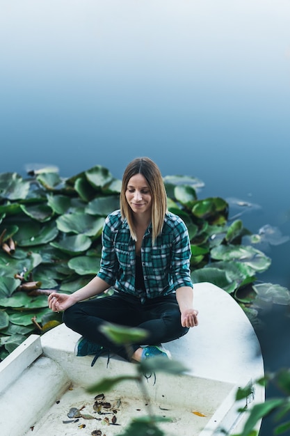 Mujer joven pacífica meditando en posición de loto sobre el agua rodeada de hojas de nenúfar